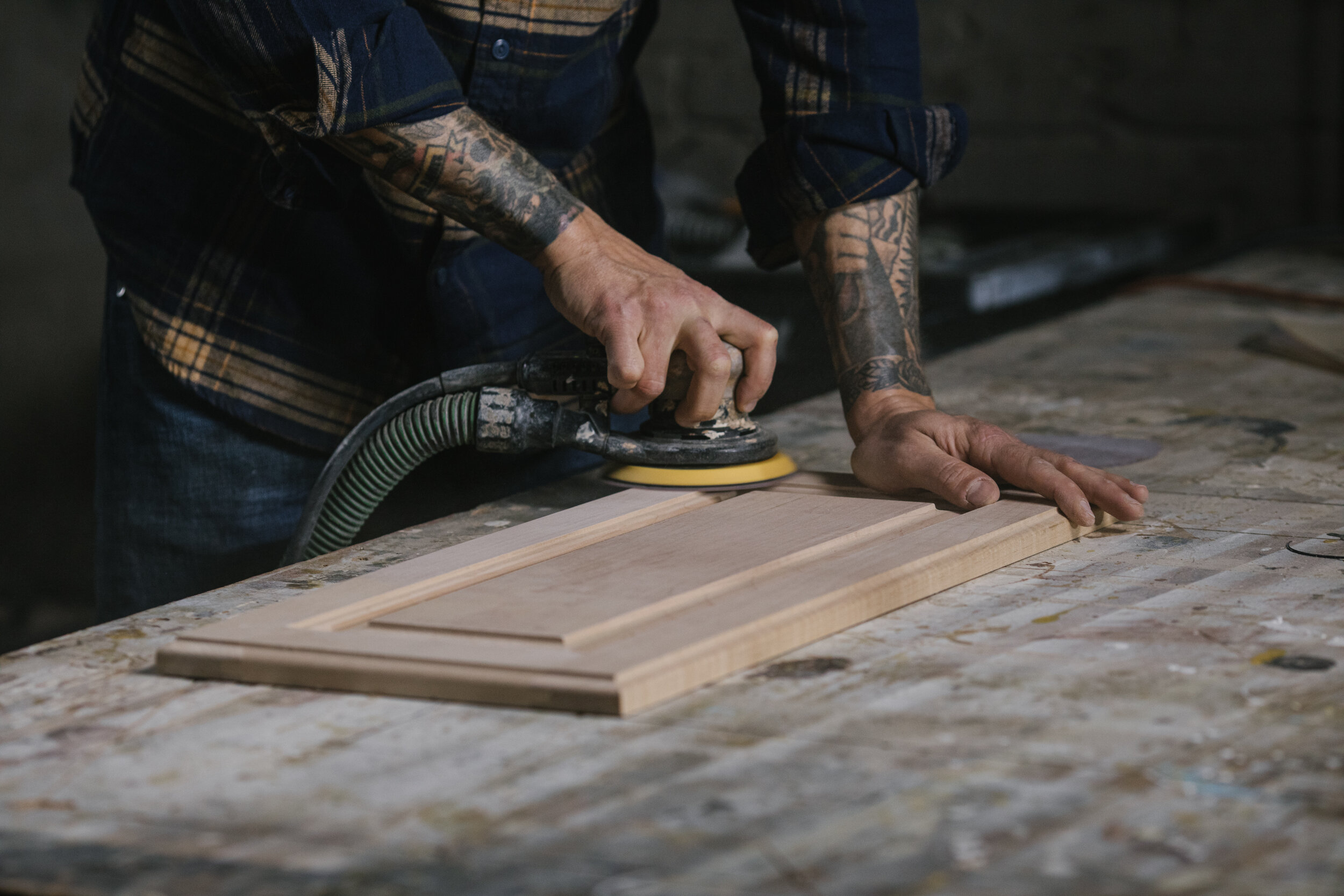 A person with tattoos on their arms using a power sander to smooth the surface of a piece of wood on a workbench in a workshop.