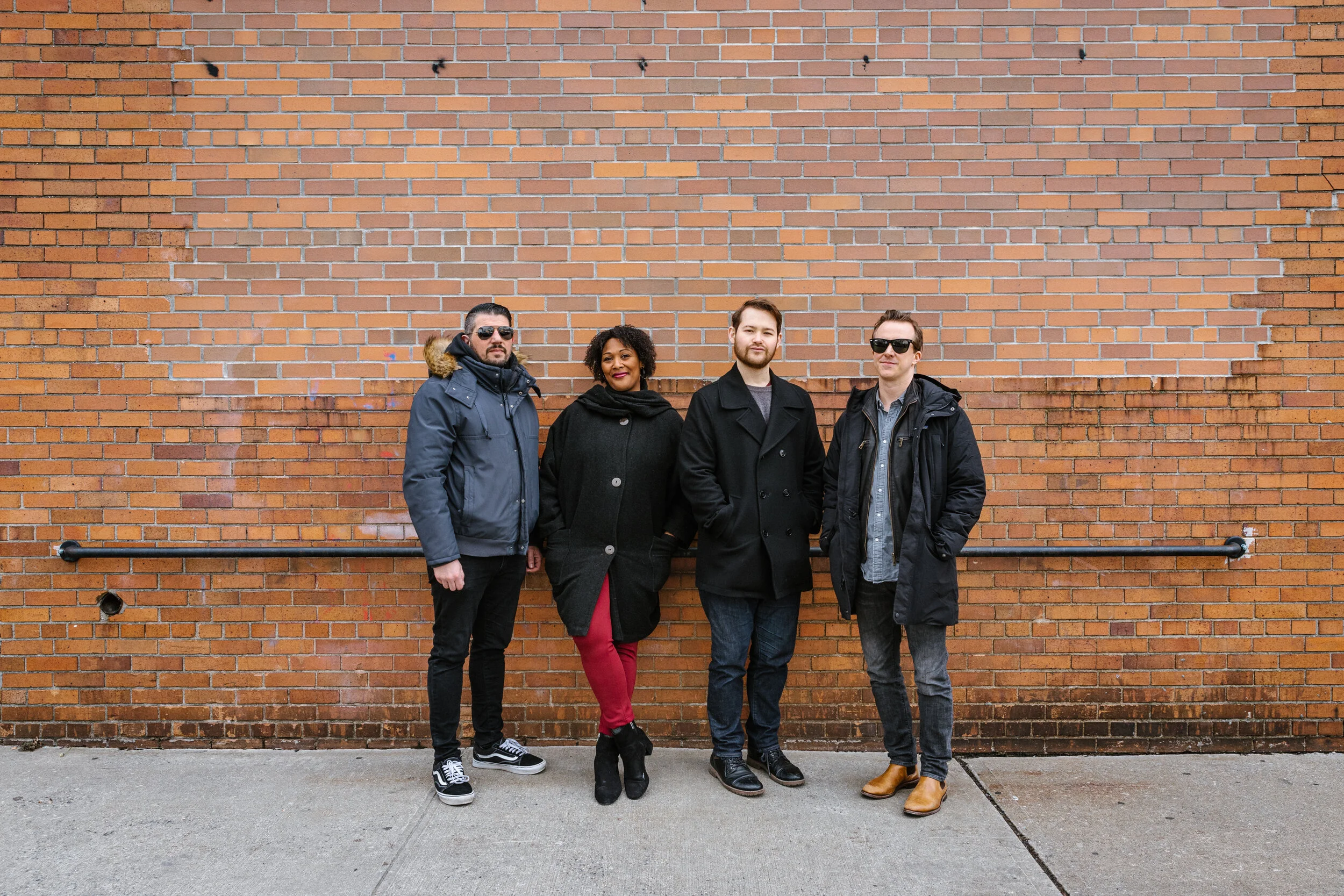 Four people, three men and one woman, standing against a brick wall outdoors. They are dressed in casual winter clothing, with the two men on the ends wearing sunglasses and jackets. The woman is in the middle, wearing a black coat and red pants, and is standing with one foot crossed in front of the other. They are posing confidently for the photo.