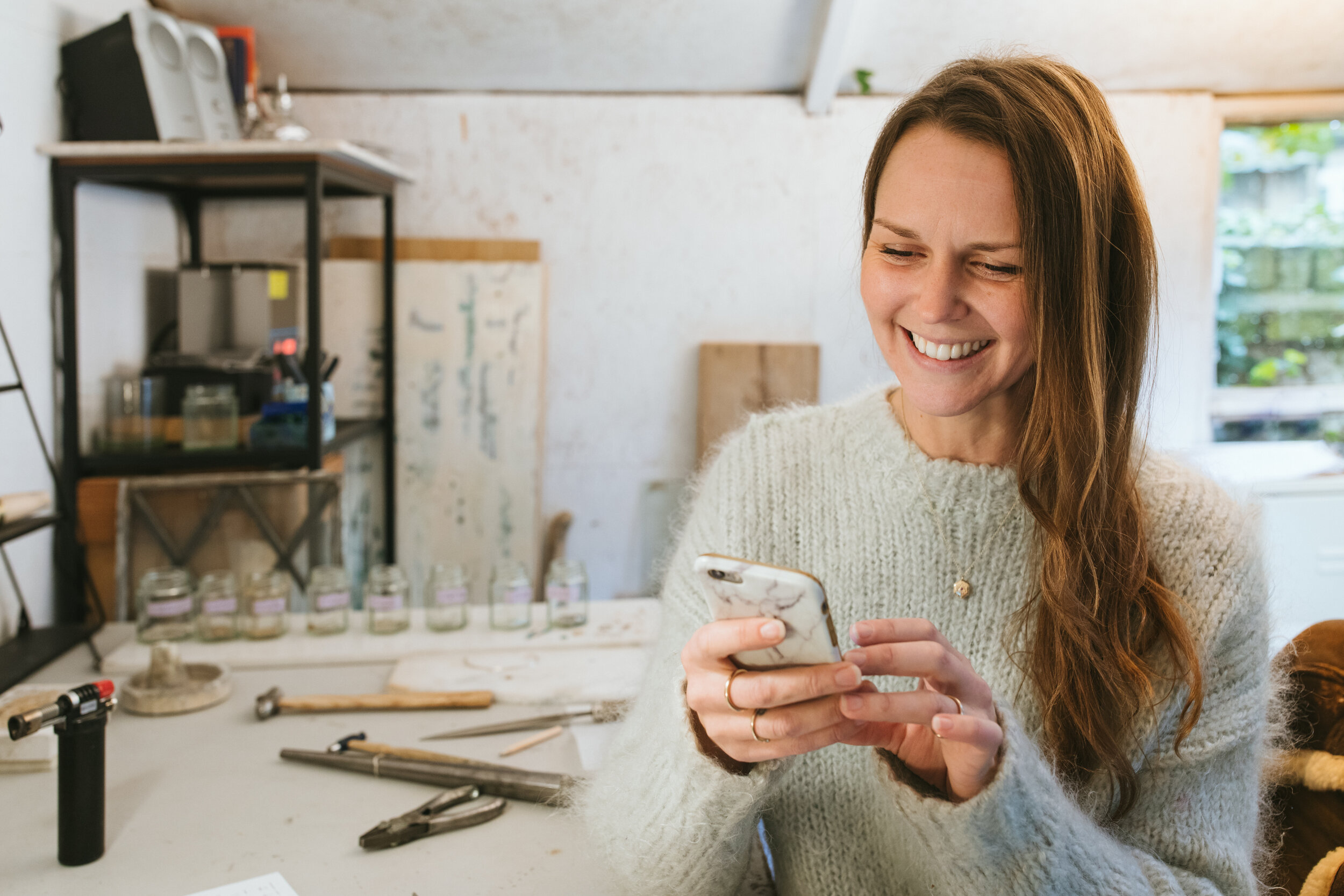 A woman with long brown hair smiling while looking at her phone, in a workshop with tools and jars in the background.
