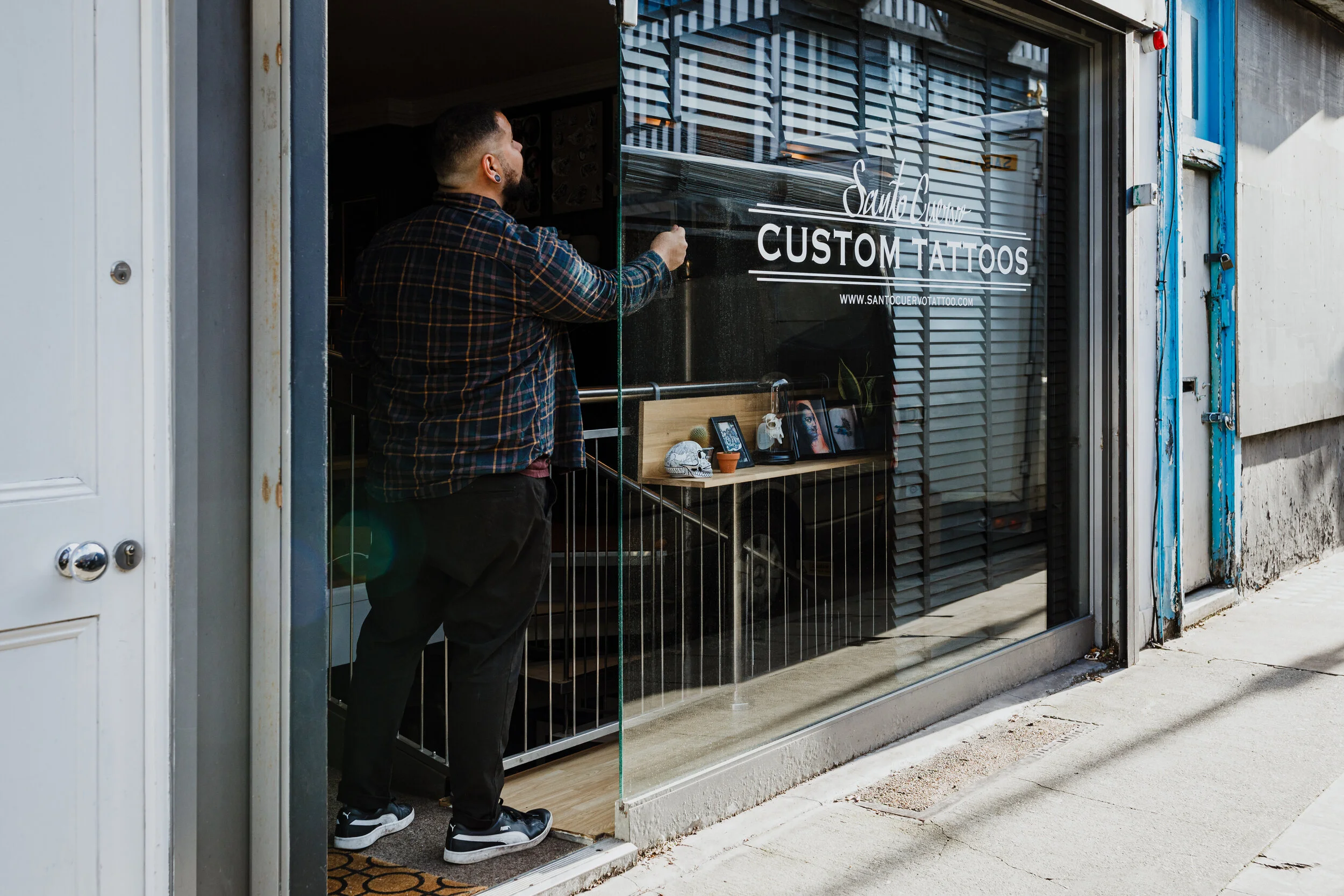 A man with tattoos and earrings is applying a decal to the glass door of a tattoo studio called 'Santo Cuervo Custom Tattoos.'