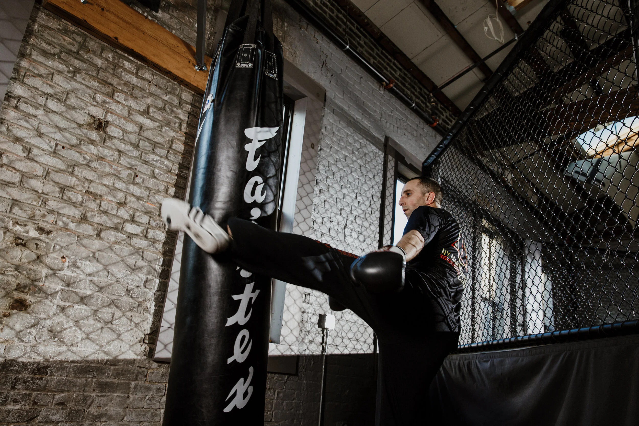 A man training in a gym, kicking a black boxing heavy bag with white text, against an industrial-style brick wall and a chain-link fence.