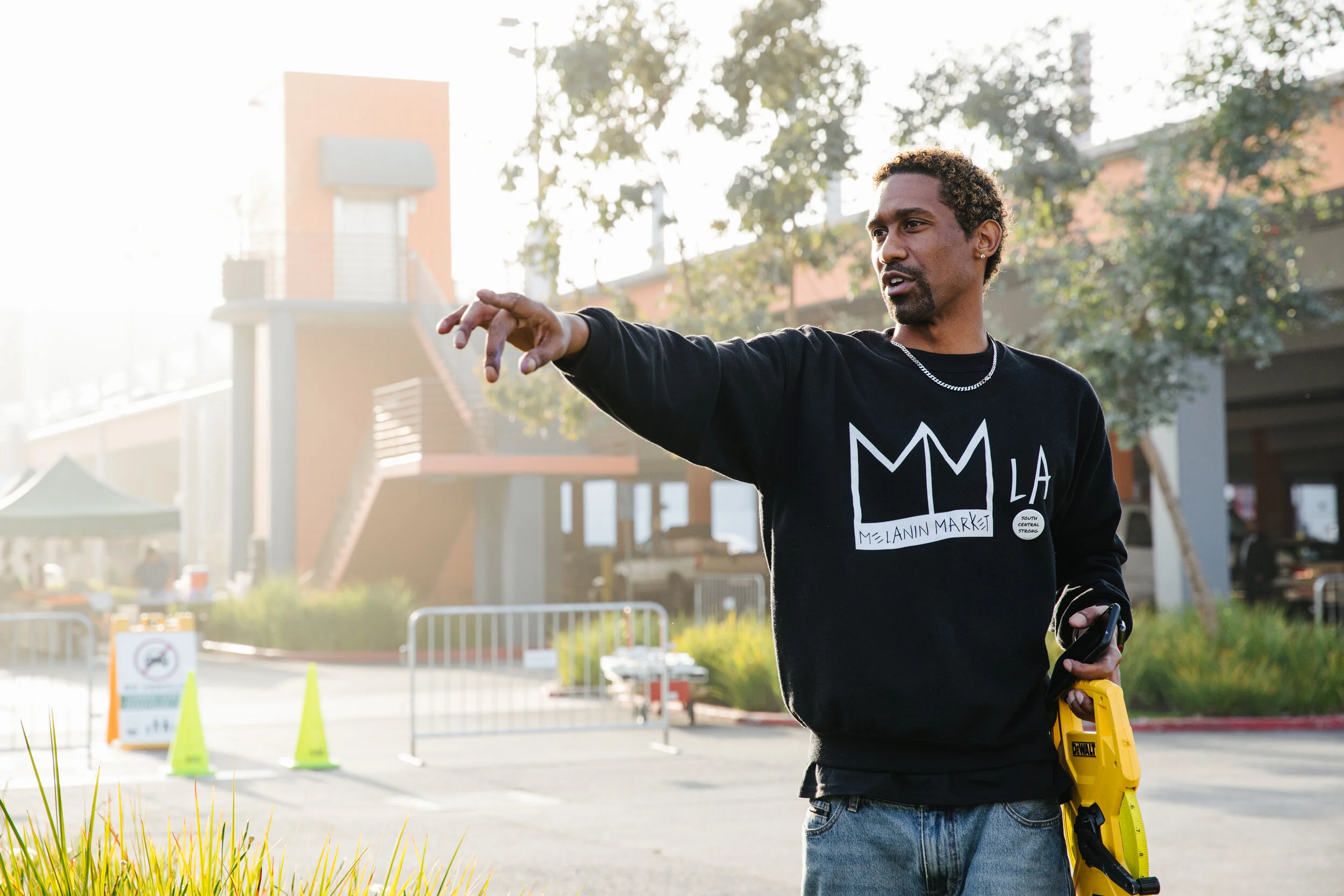 A man with curly hair and earrings is pointing while holding a laser measuring device in an outdoor setting with trees and buildings.