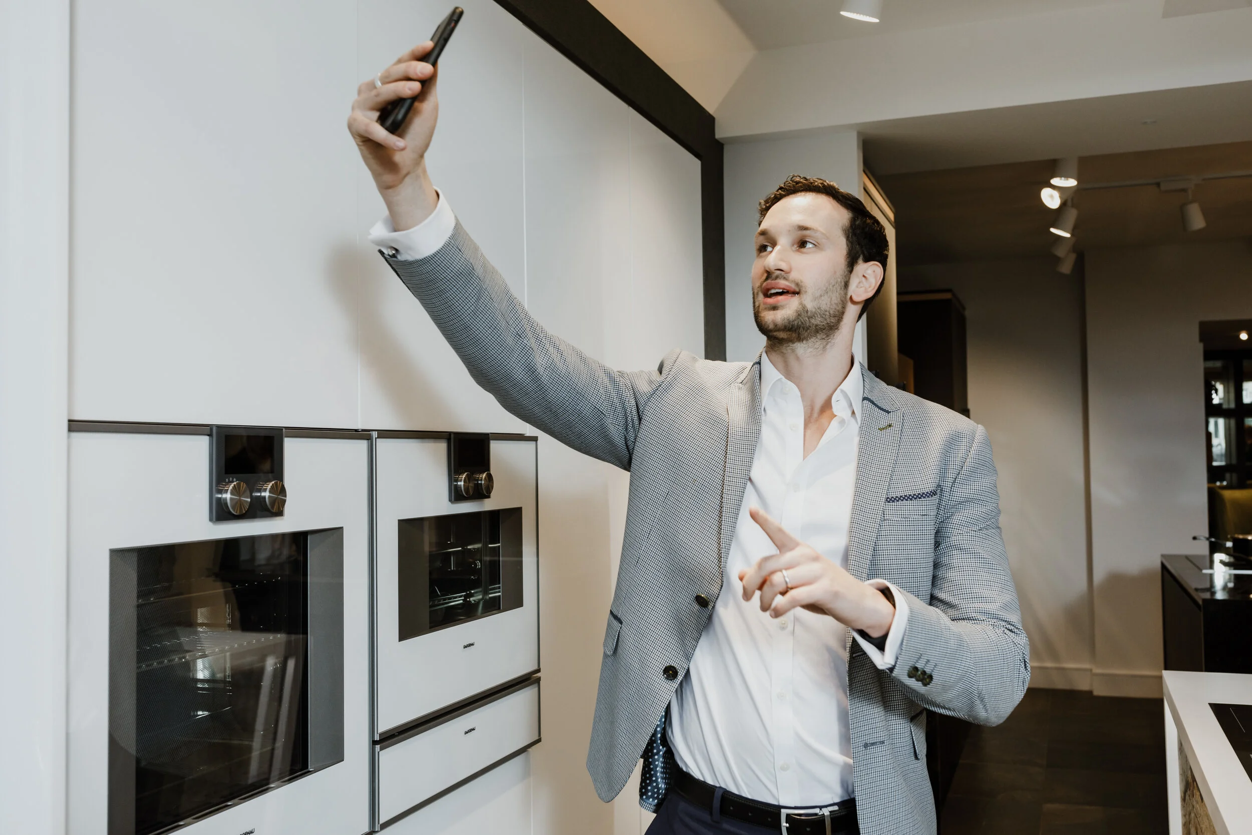 A man in a grey checkered suit is taking a selfie in what appears to be a modern kitchen or showroom, with white cabinets and built-in appliances in the background.