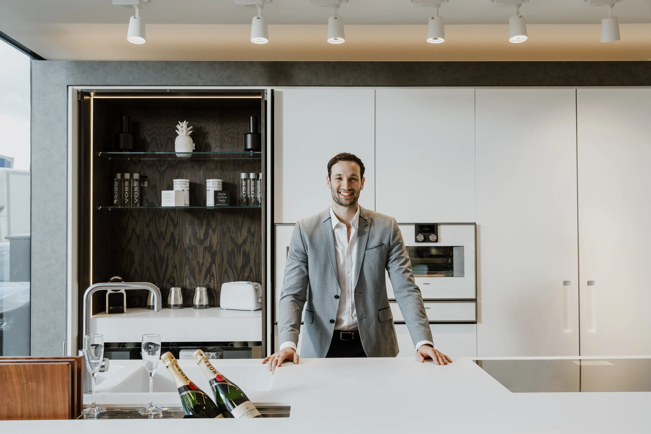 A man in a suit smiling and leaning on a kitchen island in a modern kitchen with white cabinets, a black backsplash, and a glass shelf with decorative objects. There are champagne bottles, glasses, and a corkscrew on the breakfast bar.
