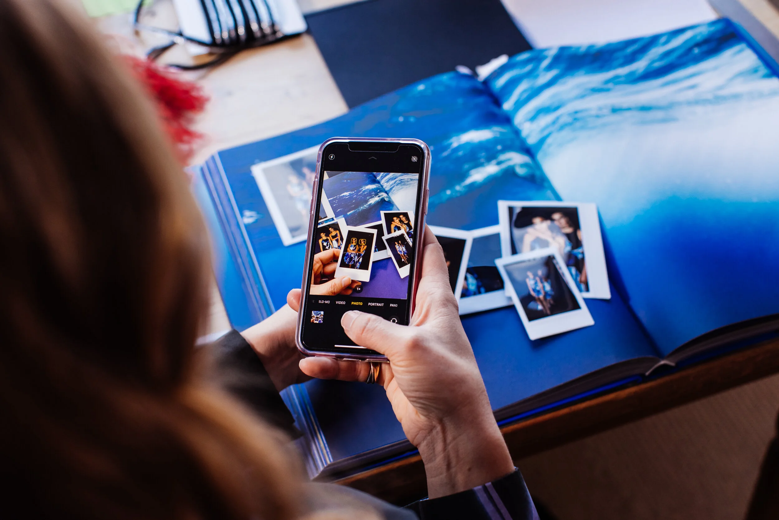 Person taking a photograph of printed photographs or snapshots with a smartphone, with a photo album or scrapbook open on a blue surface in the background.