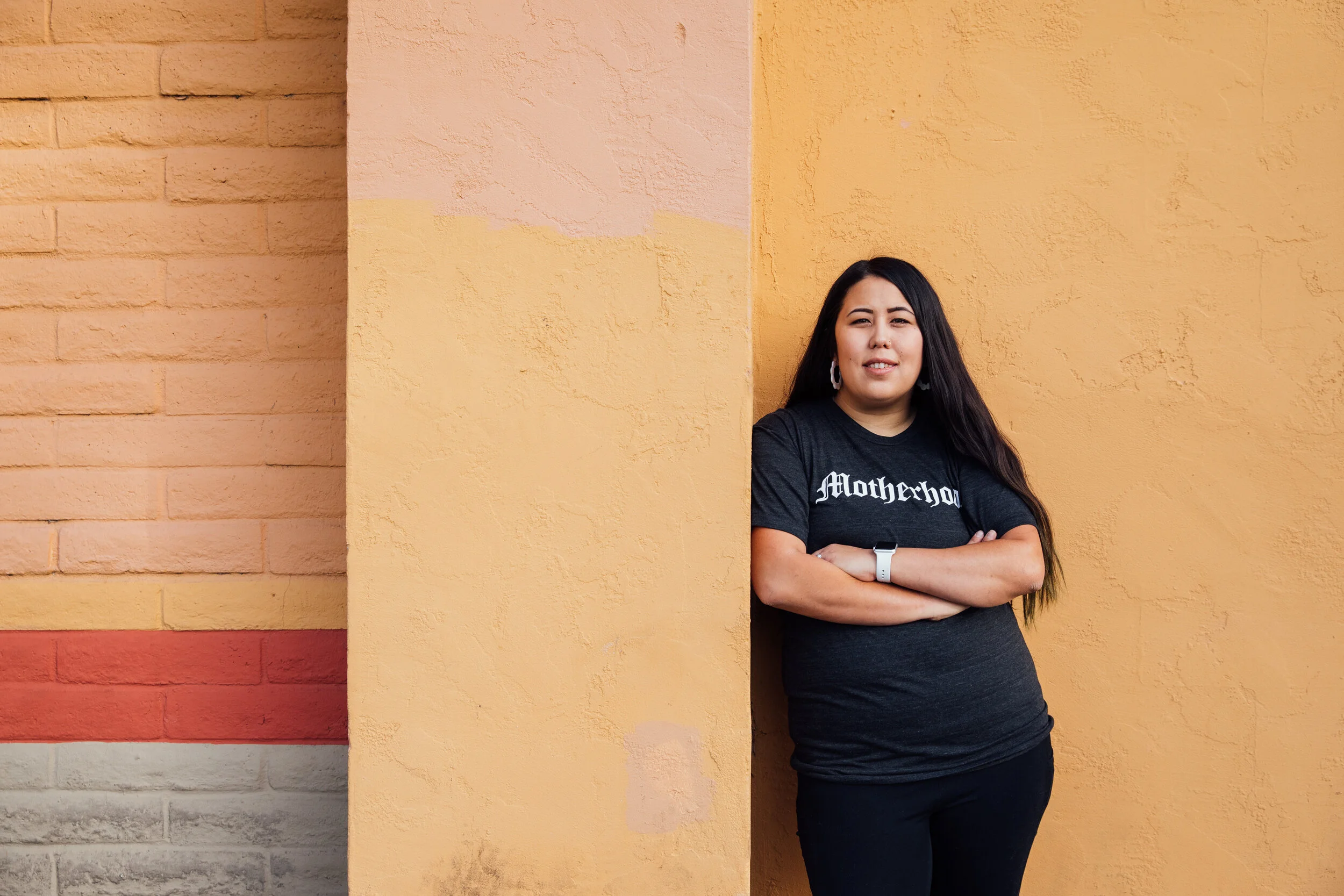 A woman with long dark hair stands with arms crossed, leaning against a yellow wall, wearing a black T-shirt and earrings.