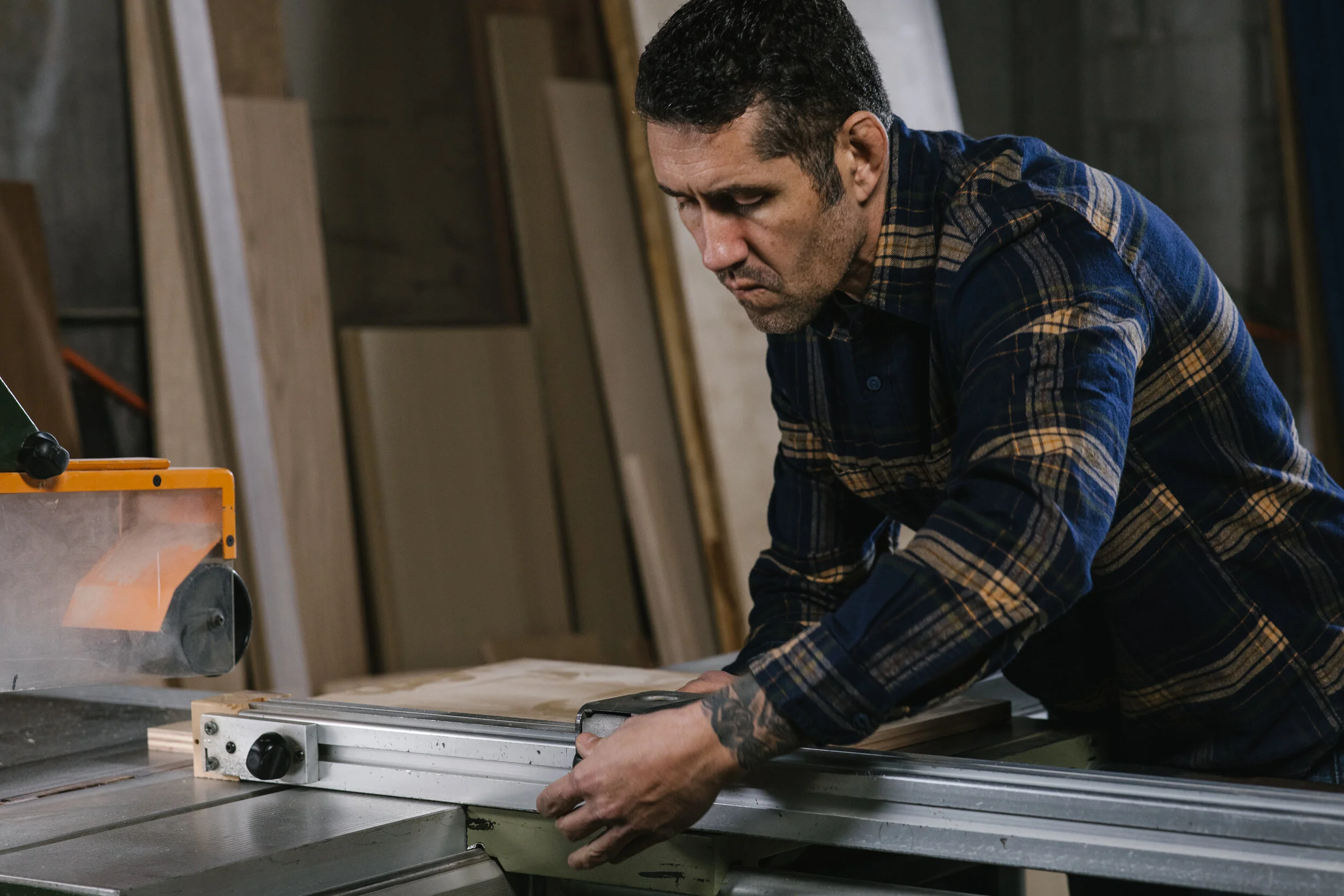 A man working in a woodworking shop, measuring a piece of wood with a metal ruler, surrounded by wooden boards and tools.