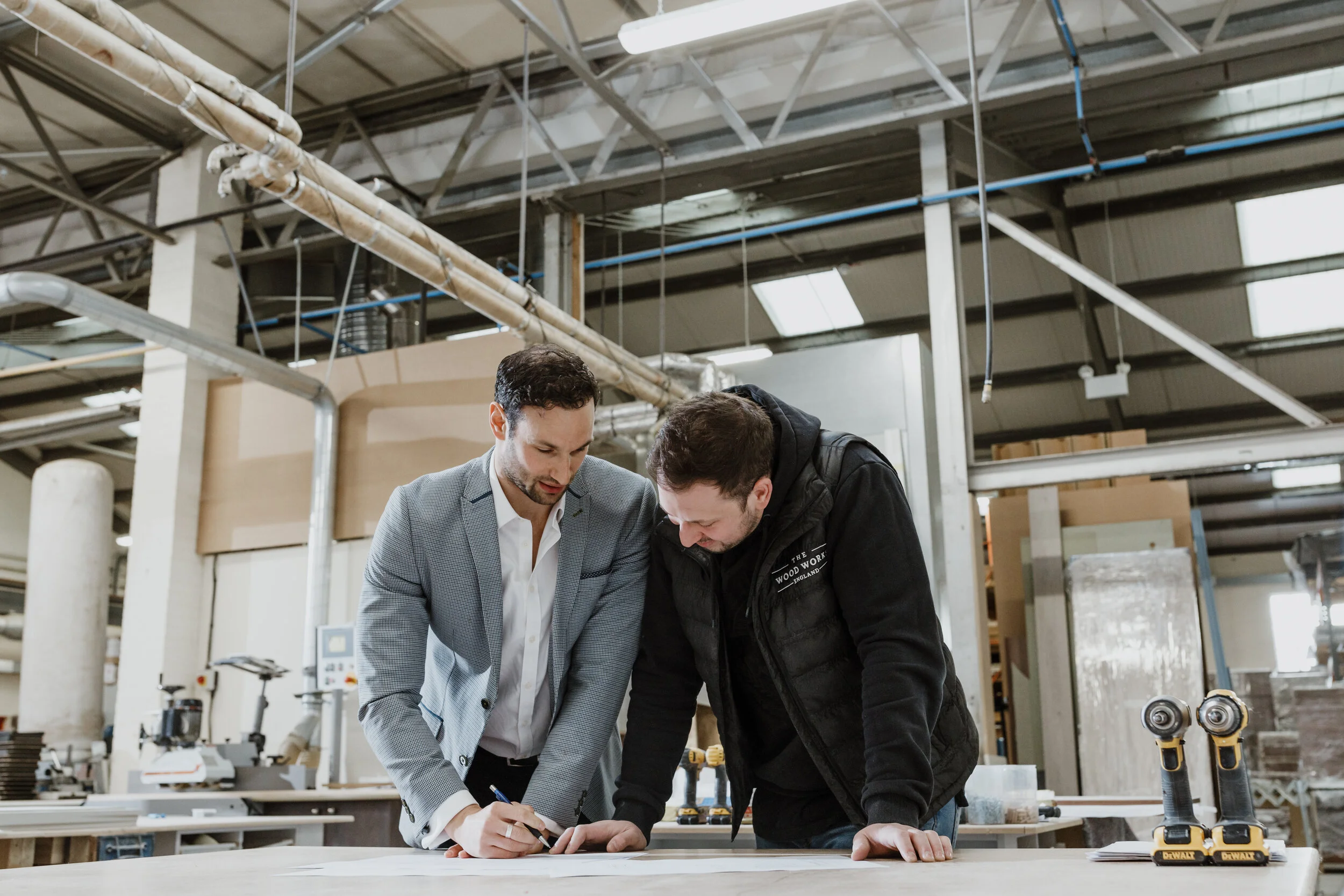 Two men are examining a piece of paper or a blueprint on a worktable in a workshop or factory. One man is in a light suit, and the other is in a black hoodie. The workspace has various tools and equipment, and the ceiling has visible pipes and beams.