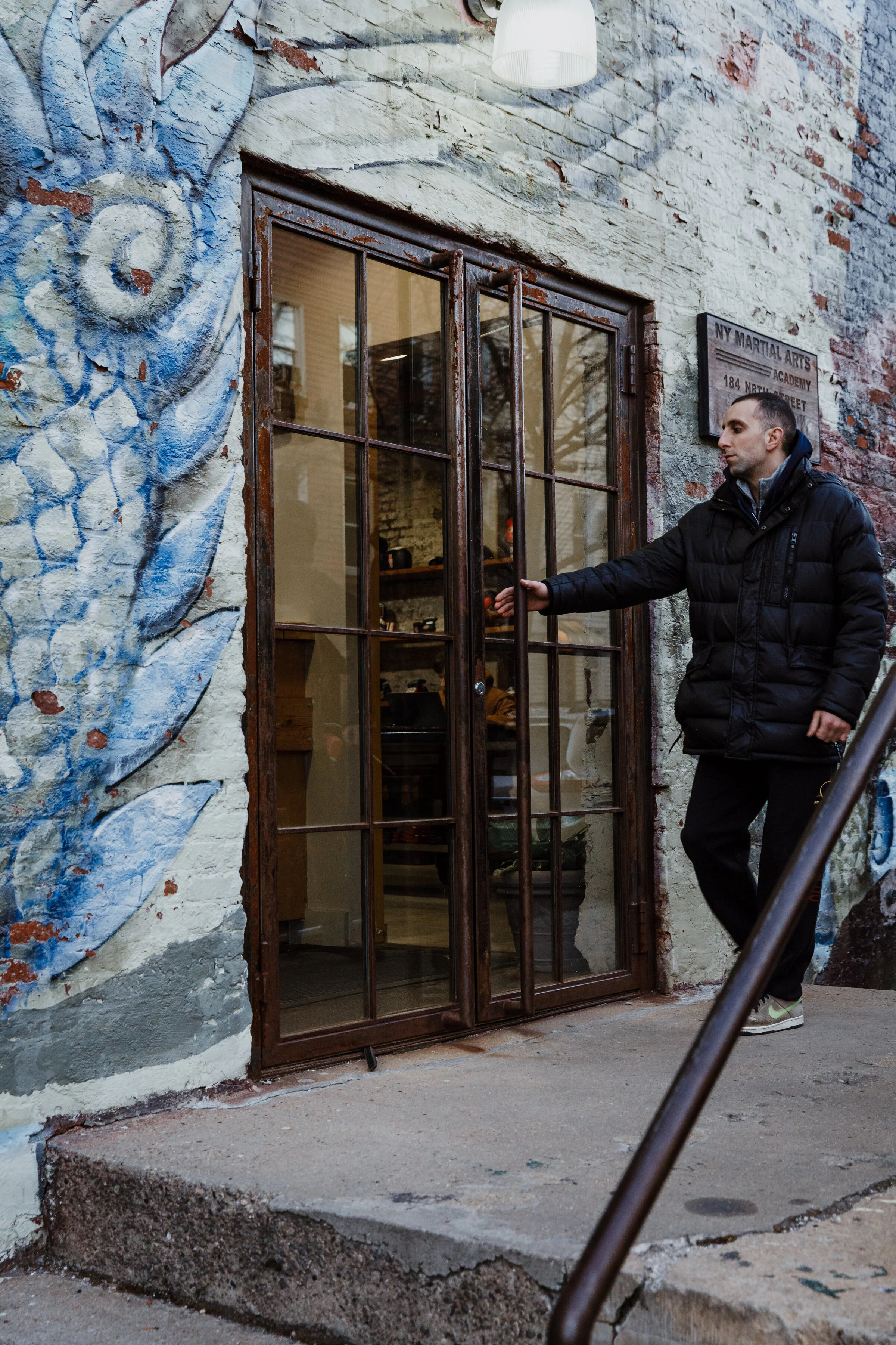 A man in a black jacket and gray sneakers standing outside a building with a painted brick wall, operating a metal-framed glass door. The wall has artwork with blue and white swirls, and a sign reads 'NY Martial Arts Academy, 184 N 6th Street'.