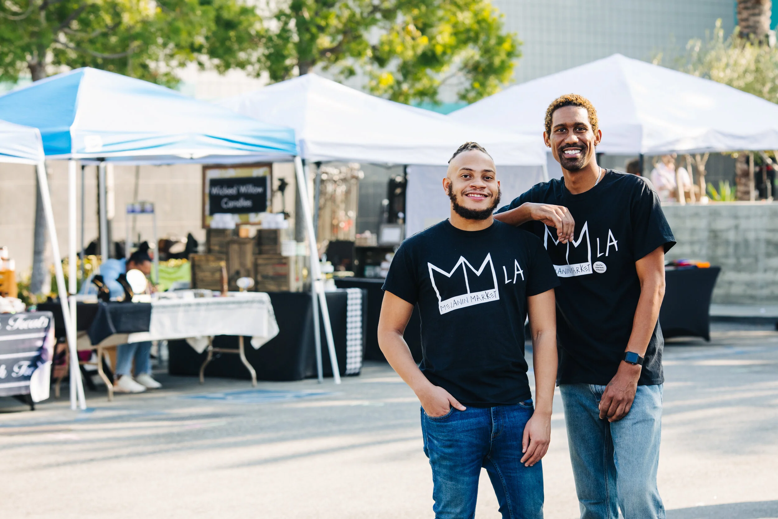 Two men smiling at a street market with vendor tents in the background