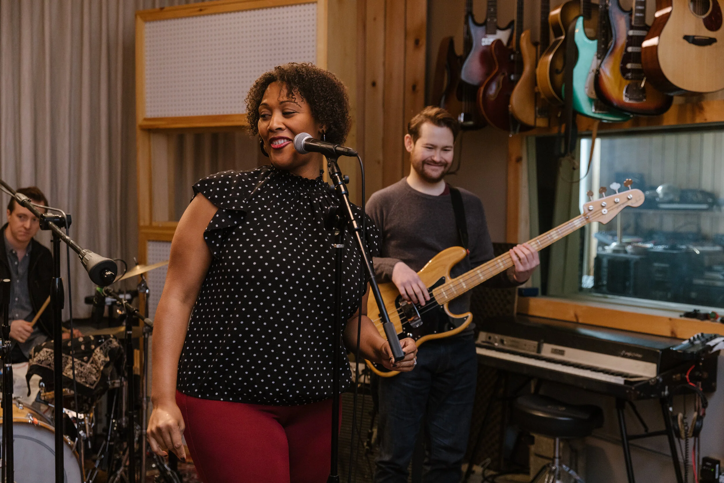 A woman singing into a microphone and a man playing bass guitar in a music studio.