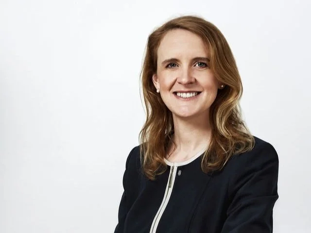 A woman with shoulder-length brown hair, smiling, wearing a black blazer and a white top, against a plain white background.