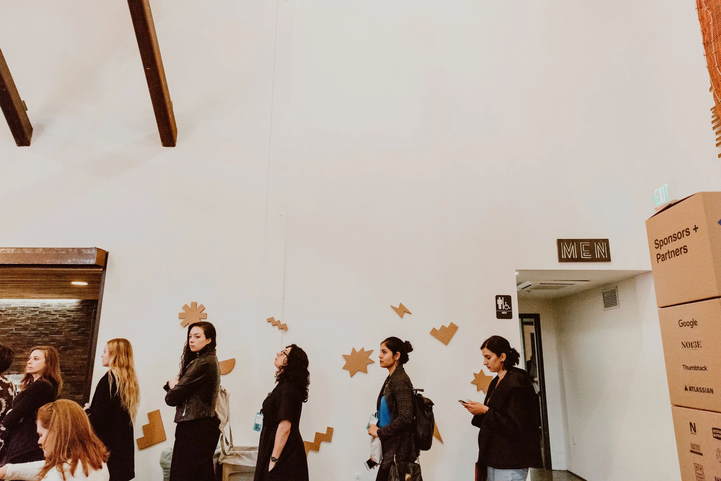 A group of women standing in a line at an indoor event, with some women wearing conference badges. The background shows a white wall with abstract orange shapes and some signage, including a men's restroom sign and informational banners.