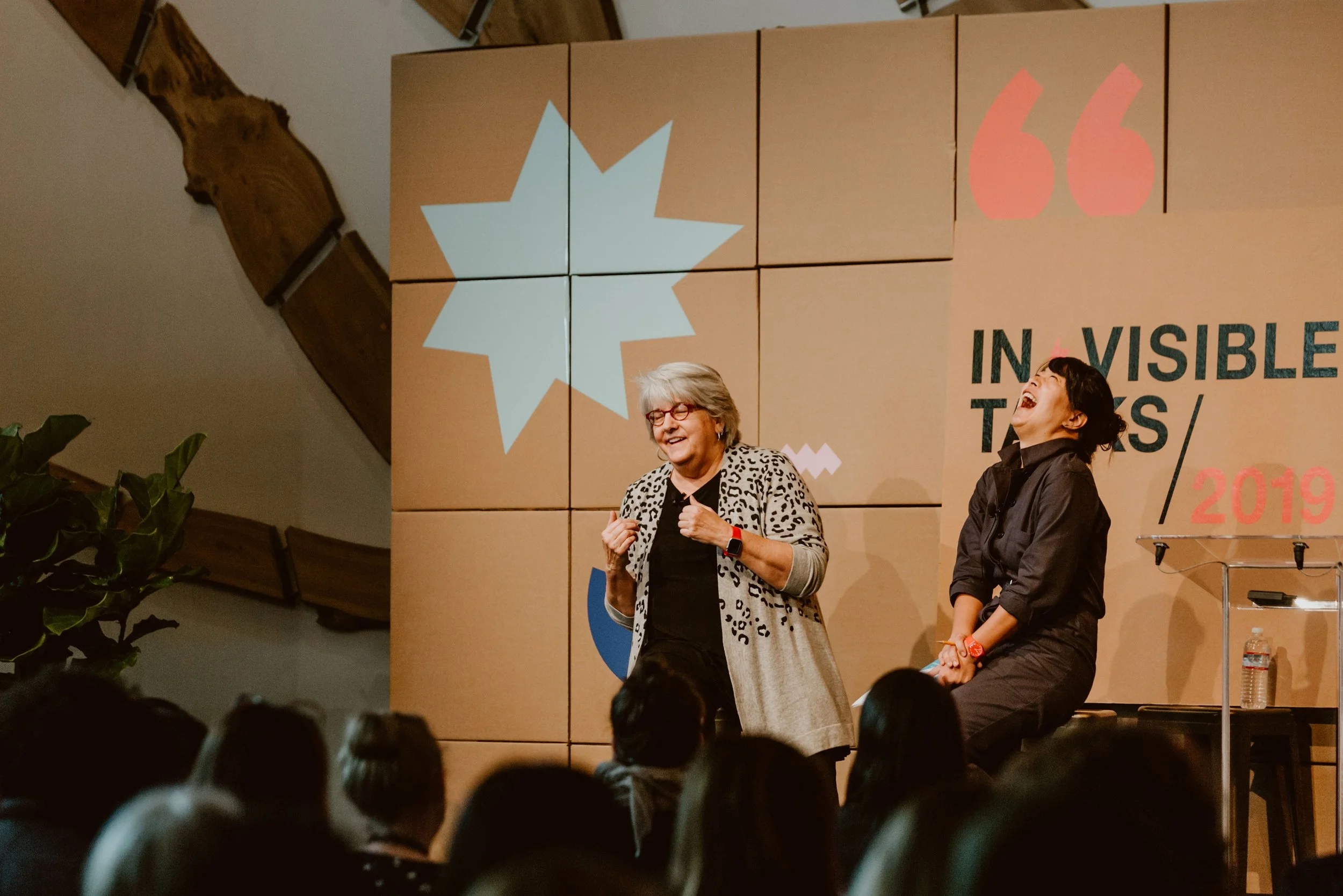 Two women speaking on stage at a conference or event with an audience. One woman is smiling and gesturing, the other is laughing. There is a large sign behind them that says 'INVISIBLE ISSUES/20'.