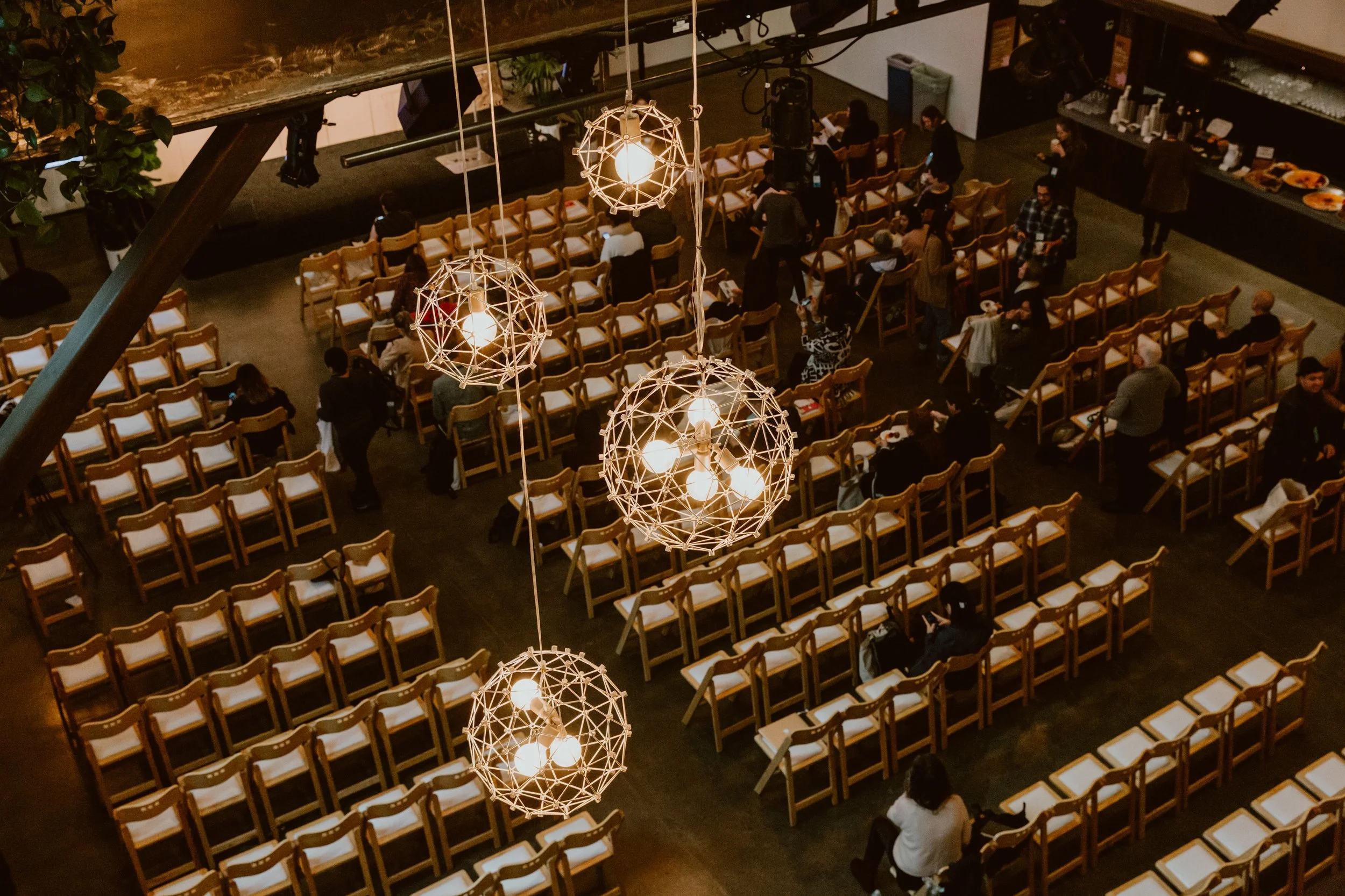 An aerial view of a large, dimly lit event space with rows of wooden chairs arranged for a gathering. Several people are seated and standing, preparing for an event. Decorative hanging light fixtures are visible in the foreground.
