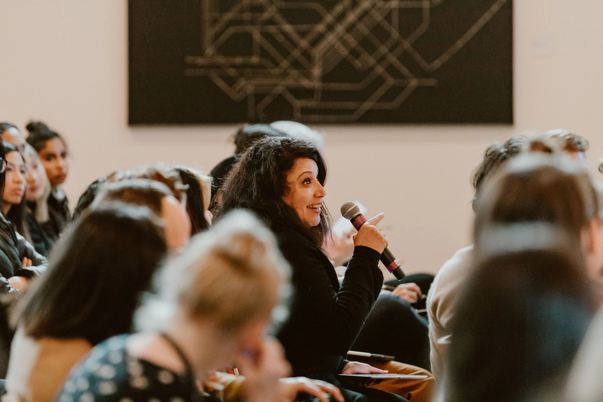 A woman with long dark hair sitting in a crowded room, holding a microphone and smiling. The room has an audience, and there is a large blackboard with a geometric pattern on the wall behind her.