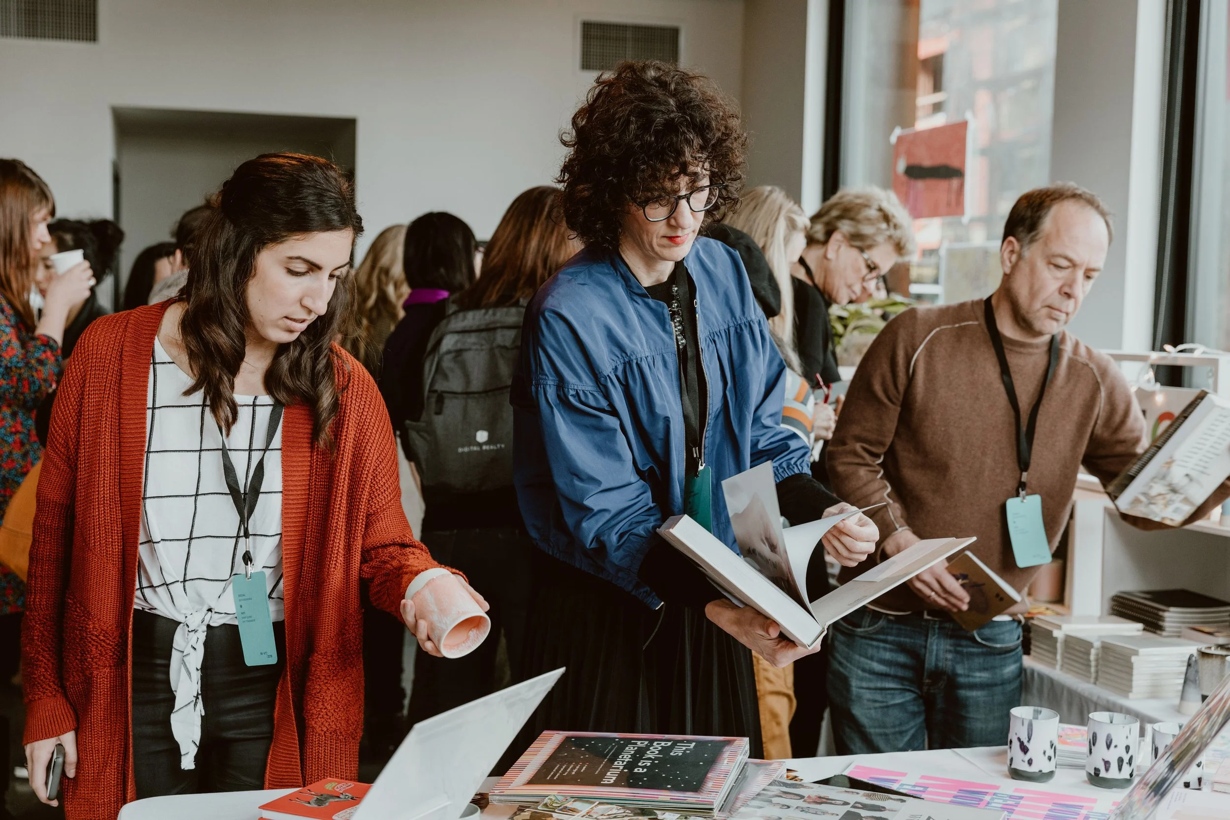 People browsing books and magazines at an indoor event or bookstore, some holding items and looking at displays.