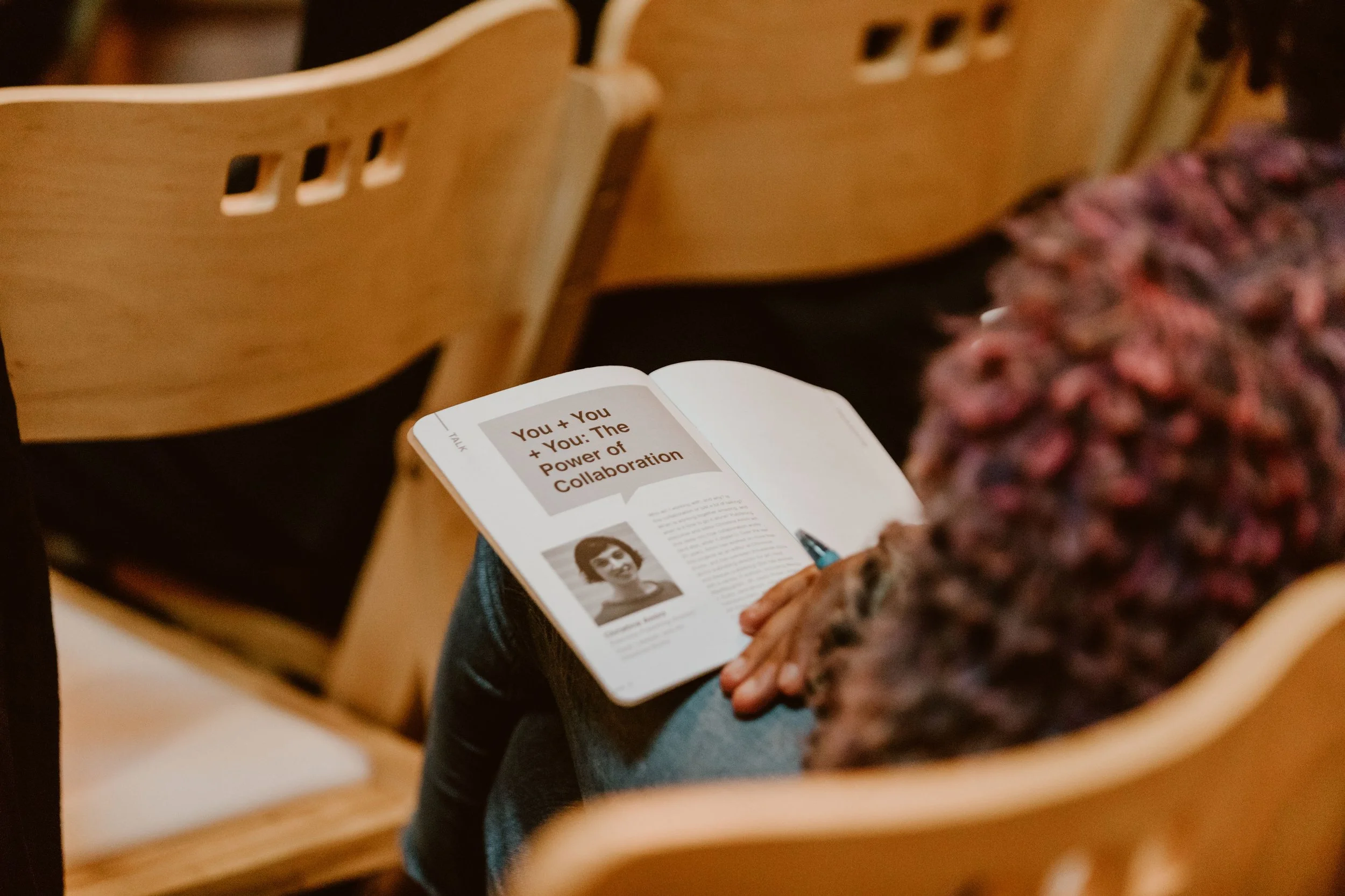 A person sitting on a wooden chair reading a magazine about collaboration, with a close-up on the magazine page showing a photo of a woman and the title 'You + You + You: The Power of Collaboration.' The person's hands are resting on their lap, and t