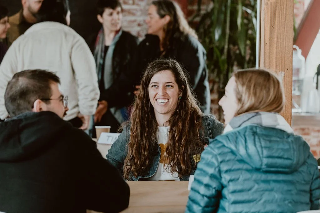 Three young women and a young man are sitting at a table in a casual restaurant or café, engaging in cheerful conversation. The woman in the center has long, wavy brown hair and is smiling, wearing a denim jacket. The woman on the right has short, li