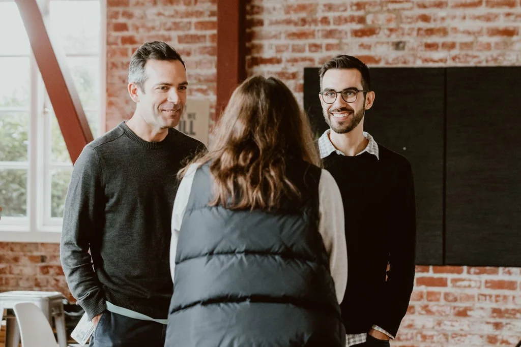 Two men smiling and a woman talking to them in a room with exposed brick wall and large window.