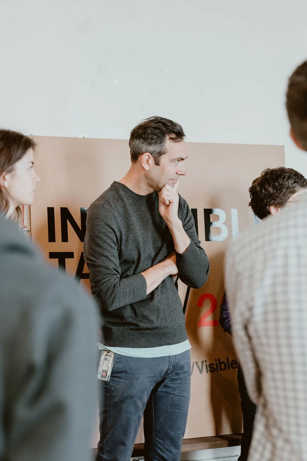 Man with dark hair and gray sweater standing in front of a beige and black sign, surrounded by other people, with his hand on his chin in thought.