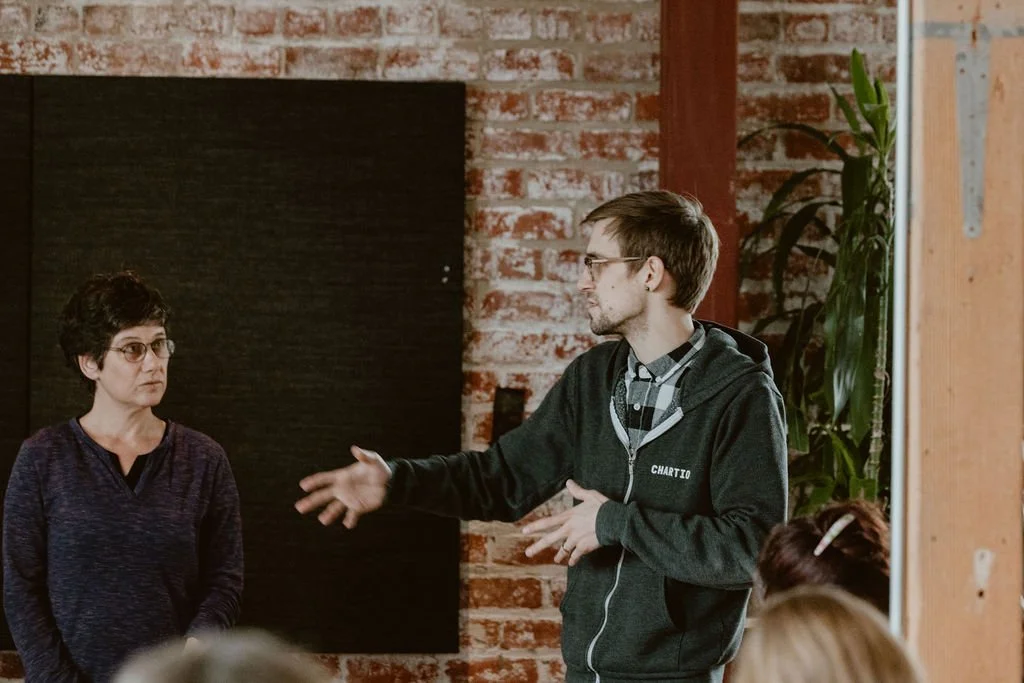 A man in a hoodie speaking and gesturing with his hand to a woman in glasses, in a room with a brick wall background.