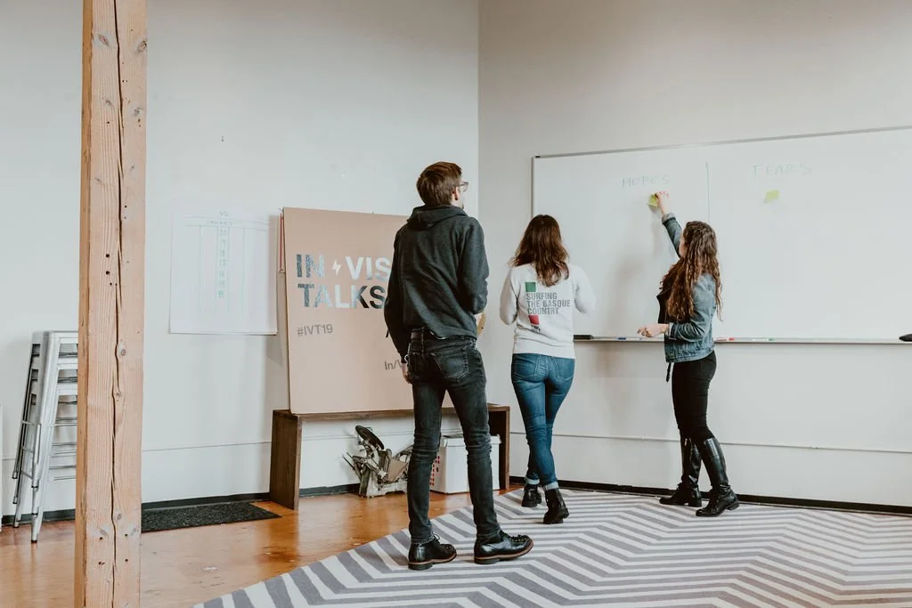 Three people standing in front of a whiteboard during a discussion or presentation in a room with wooden flooring and a striped carpet.