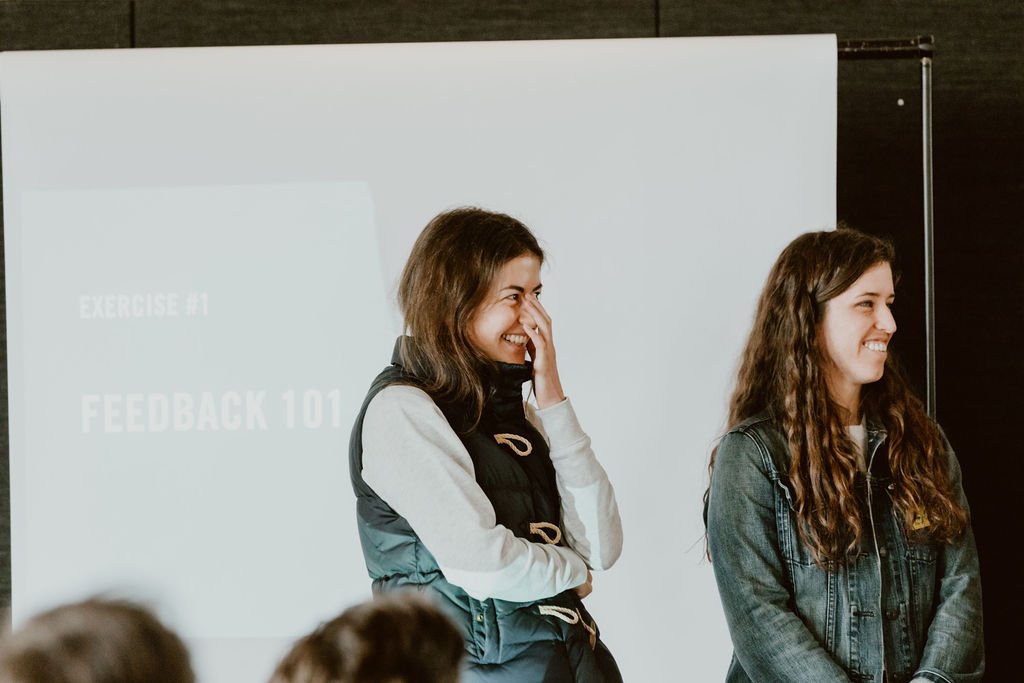 Two young women standing in a classroom or seminar setting, smiling and looking to the right, with a presentation slide in the background reading "Exercise 01" and "Feedback 101."