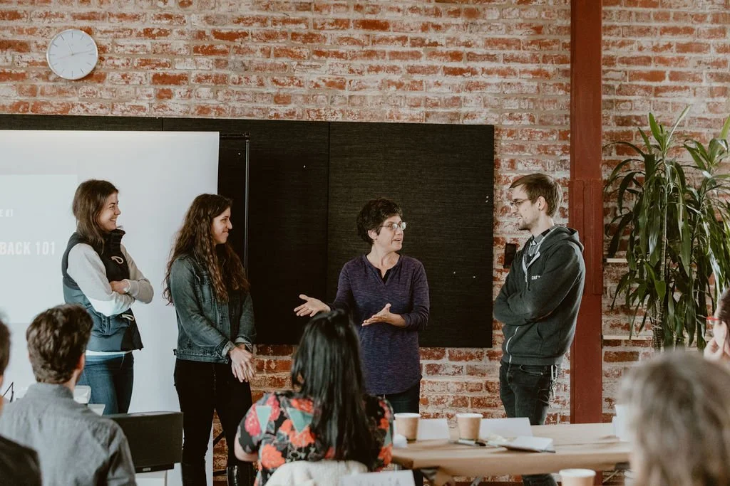 A woman is speaking to three young adults in a classroom or conference room, with some seated people and a brick wall background.