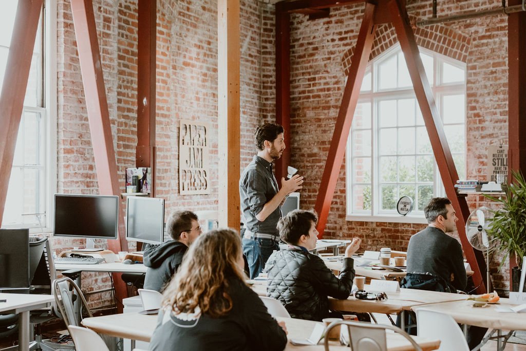 A man standing and speaking in front of a group of seated people in a bright, industrial-style office with brick walls and large windows.