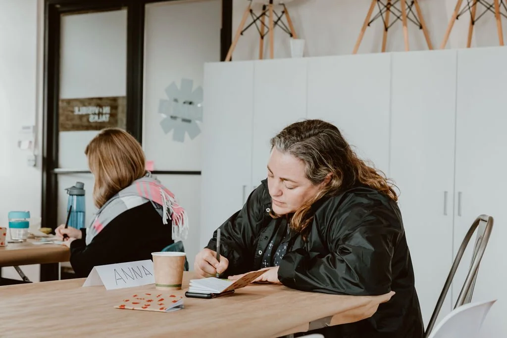 Two women sitting at a wooden table, focused on writing in notebooks during a meeting or workshop in a modern office space.
