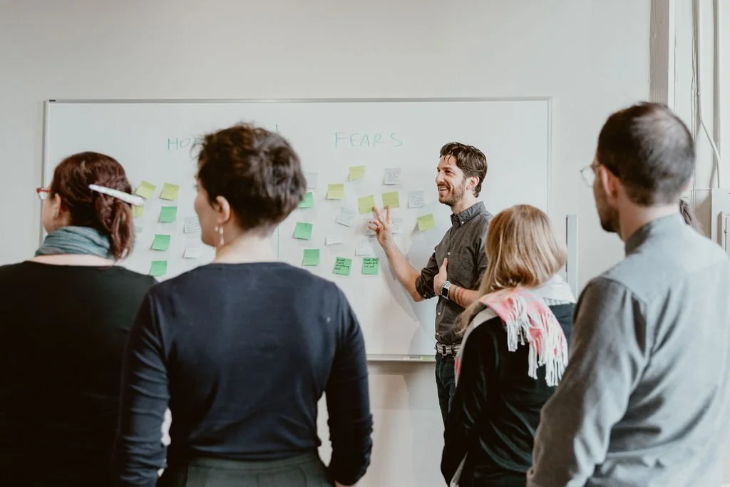 A group of people attending a presentation or workshop, listening to a man pointing at a whiteboard with notes and sticky notes.