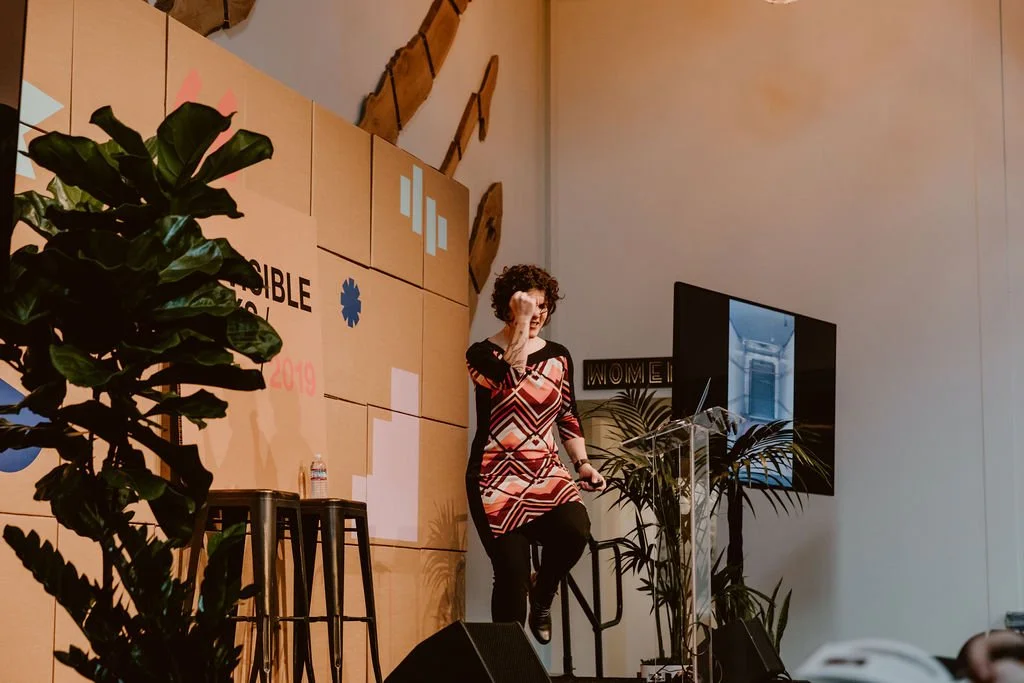 A woman with curly hair wearing a patterned dress standing on a stage with a microphone in her hand, behind her a large screen, plants, and a wall with a logo and text.