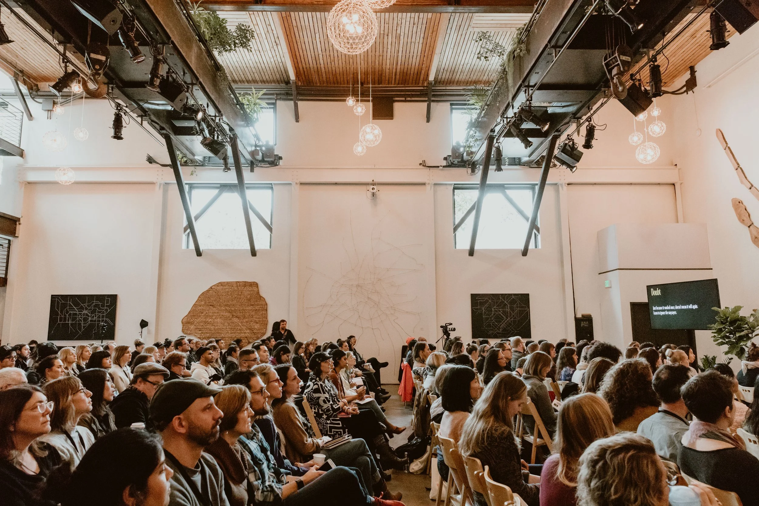A large audience seated in a conference or seminar room, facing the front. The room has high ceilings with wooden panels and decorative lighting fixtures hanging from above. There are large windows letting in natural light, and the walls are decorate