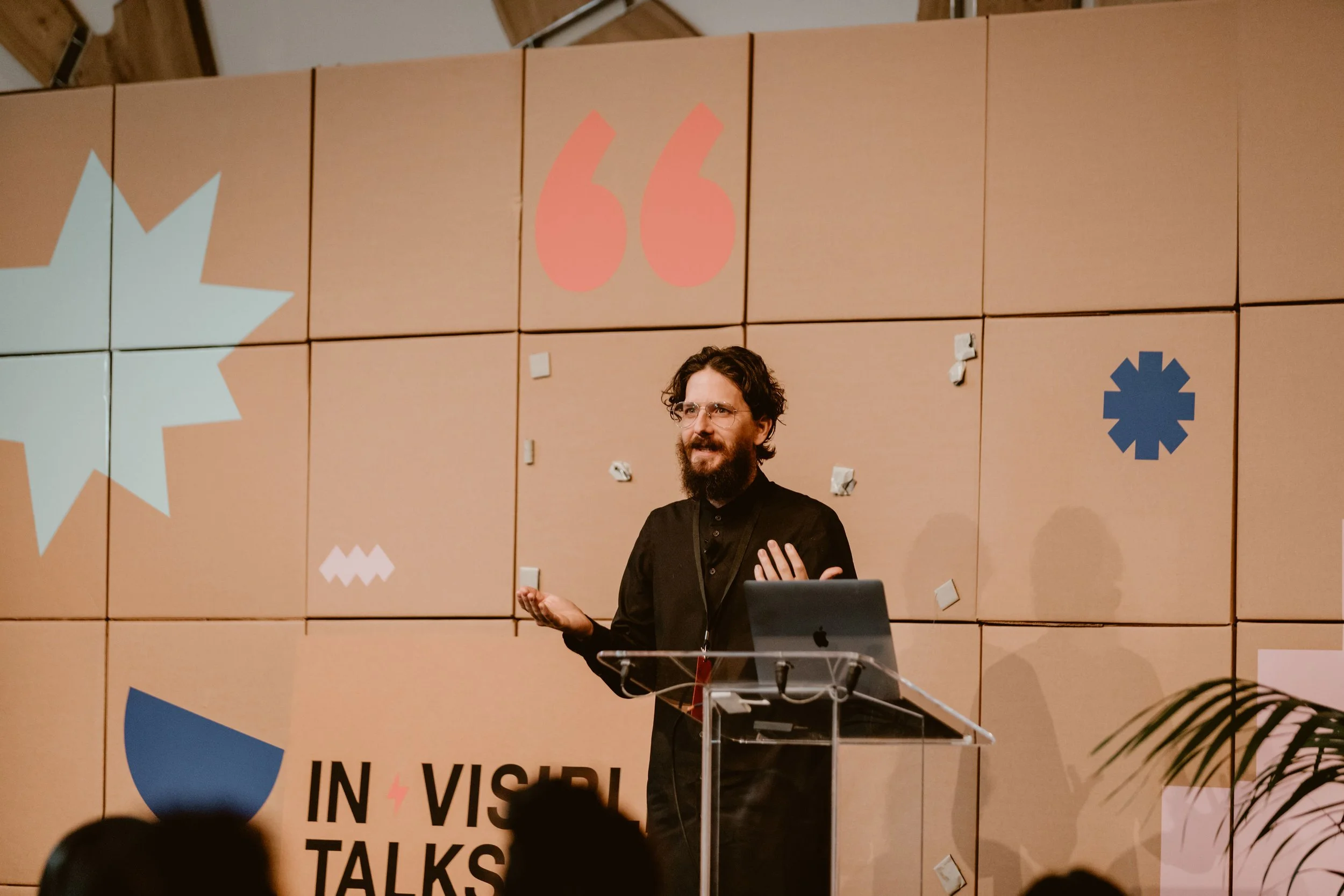 A man with glasses and a beard giving a presentation at a lectern, with a colorful background featuring various symbols and the words "INVISIBLE TALKS."