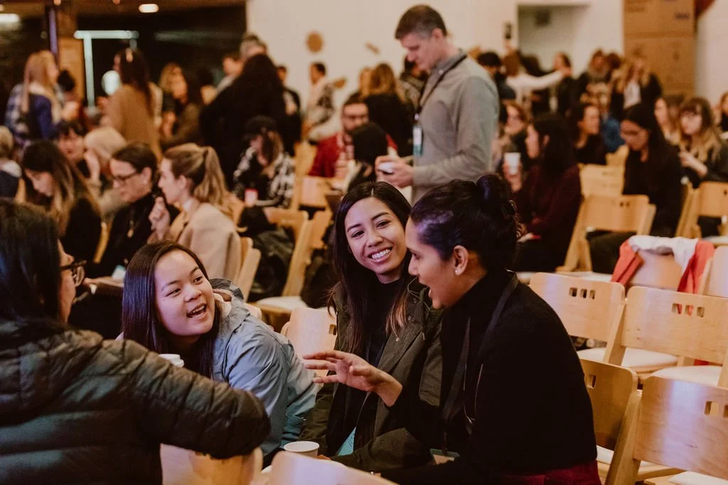 A group of four women seated and talking at a conference or event, with many other attendees in the background.