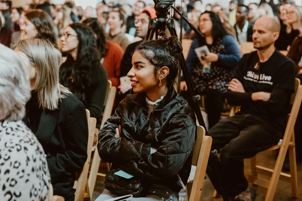 People attending an indoor event, sitting in chairs, some taking notes, with a young woman in the foreground wearing a black jacket and earrings, smiling and looking attentive.