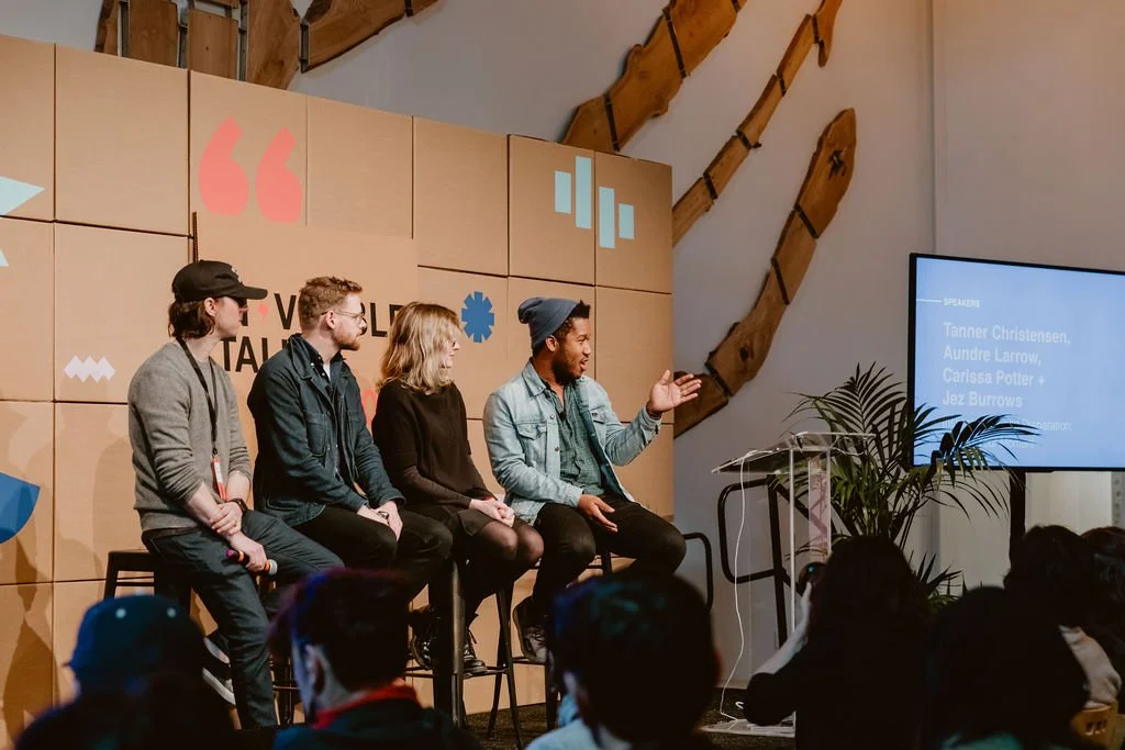 Panel discussion with five speakers sitting on high stools on stage, audience facing them, modern conference room with wooden wall art, and a large screen to the right displaying speaker names.