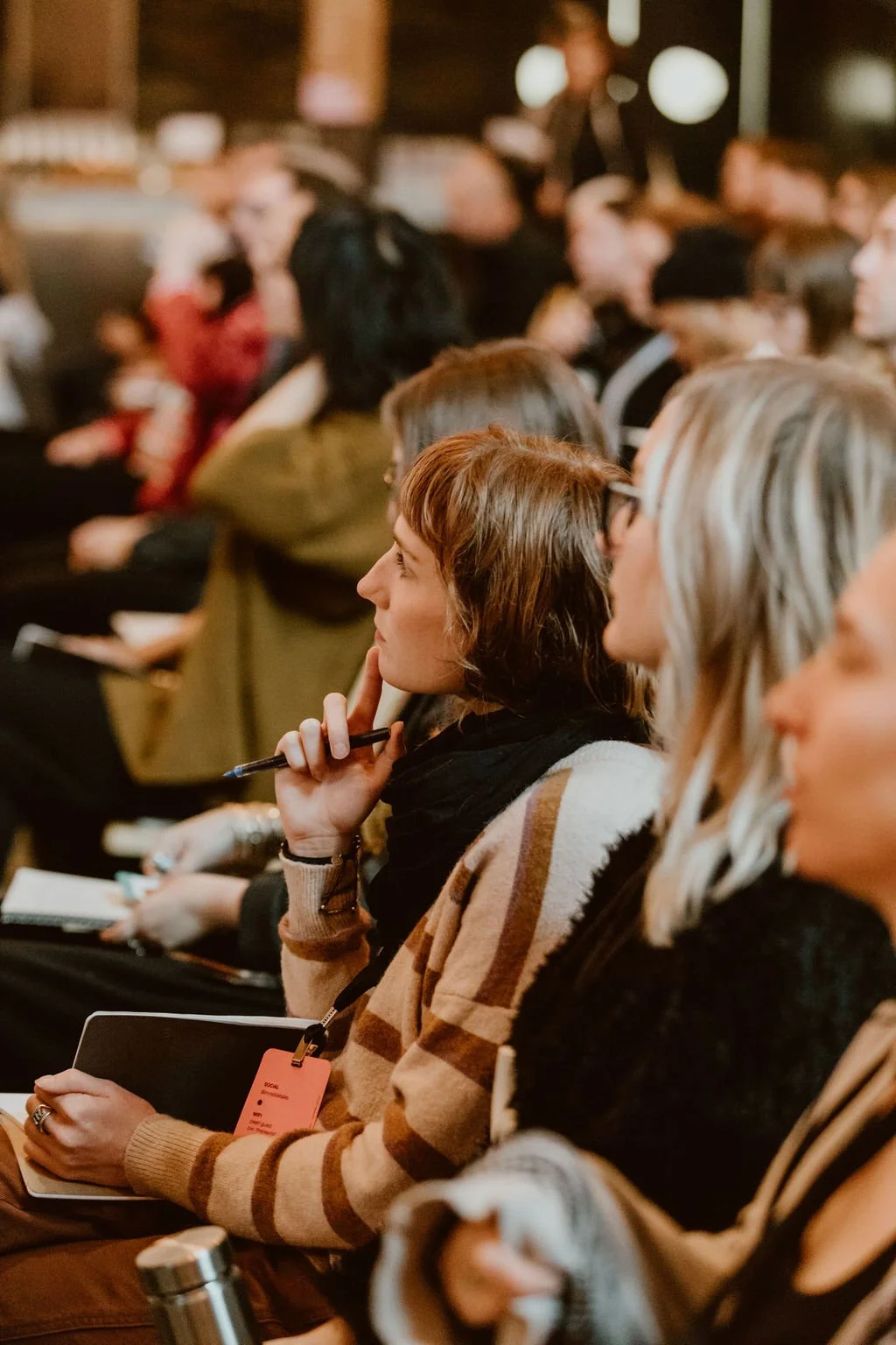 Attendees sitting and listening at a conference, some taking notes, in a dimly lit room.