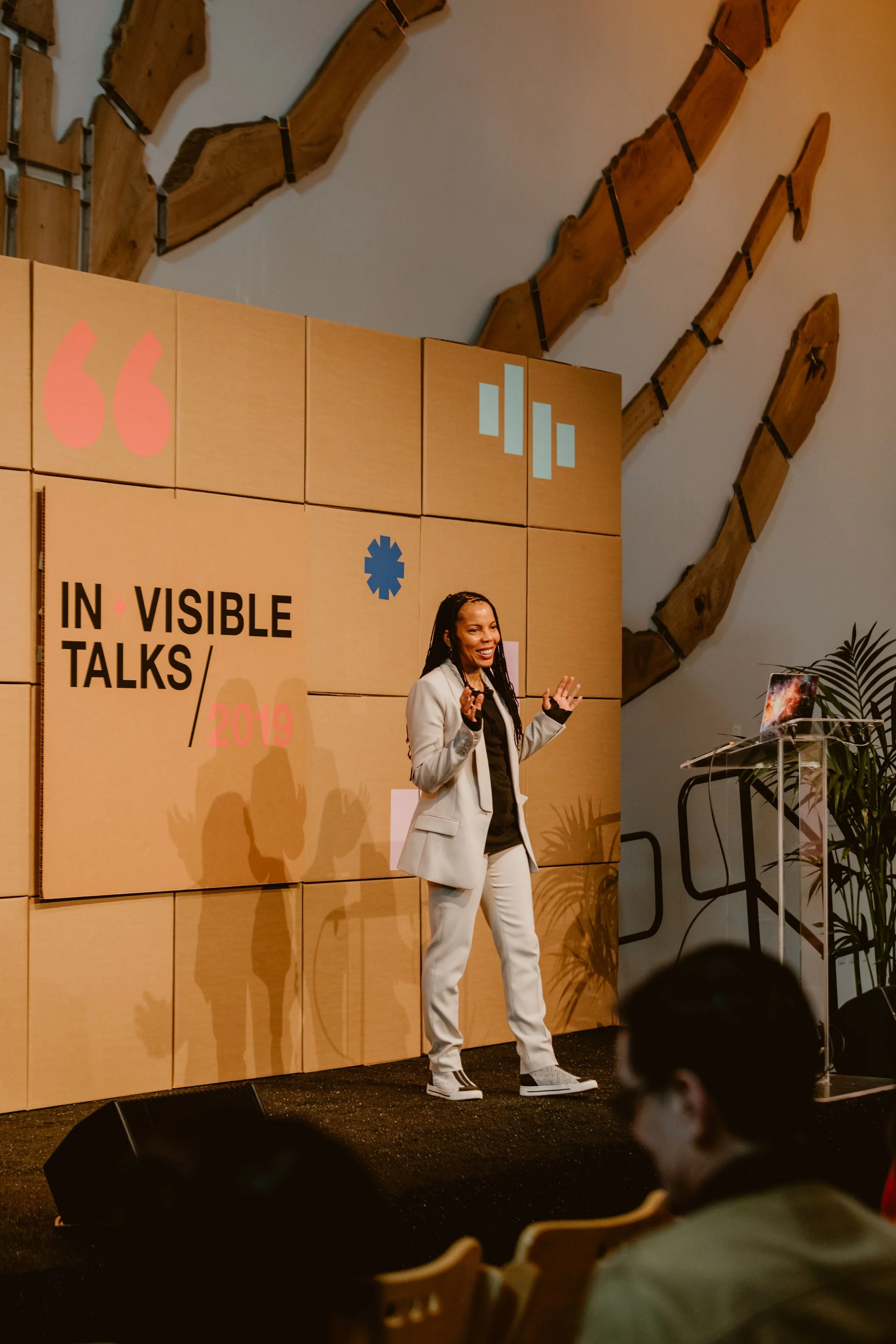 A woman in a light-colored suit speaks at a conference stage with a backdrop that reads 'INVISIBLE TALKS 2019.' She is smiling and gesturing with her hands, and there is a laptop and plant on a table beside her. Audience members are visible in the fo