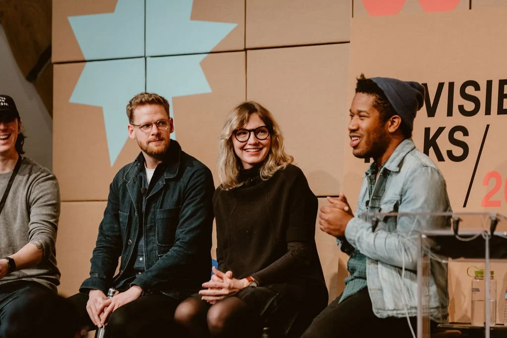 Three people sitting and smiling during a panel discussion or event, with a large logo or design projected on the wall behind them.