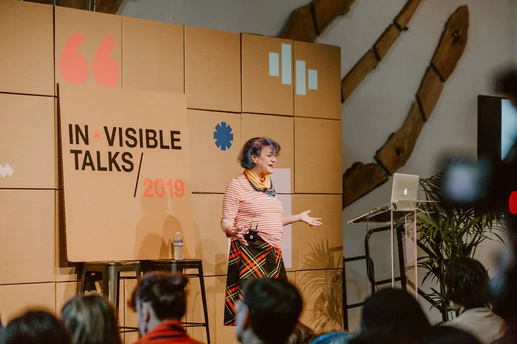 A woman giving a talk at an event called 'Invisible Talks 2019'. She is standing on stage with a microphone in her hand, wearing a striped top and colorful skirt. There is a laptop on a stand nearby, and a large clock in the background with an audien