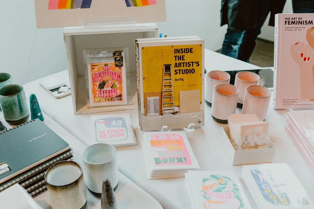 A display table with books, notebooks, cups, and decorative items at an art or book event.