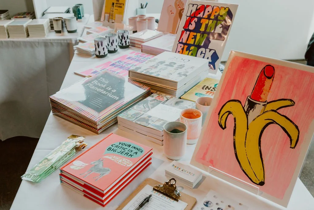 Display table with colorful books, posters, cups, and art supplies at an art or craft fair.