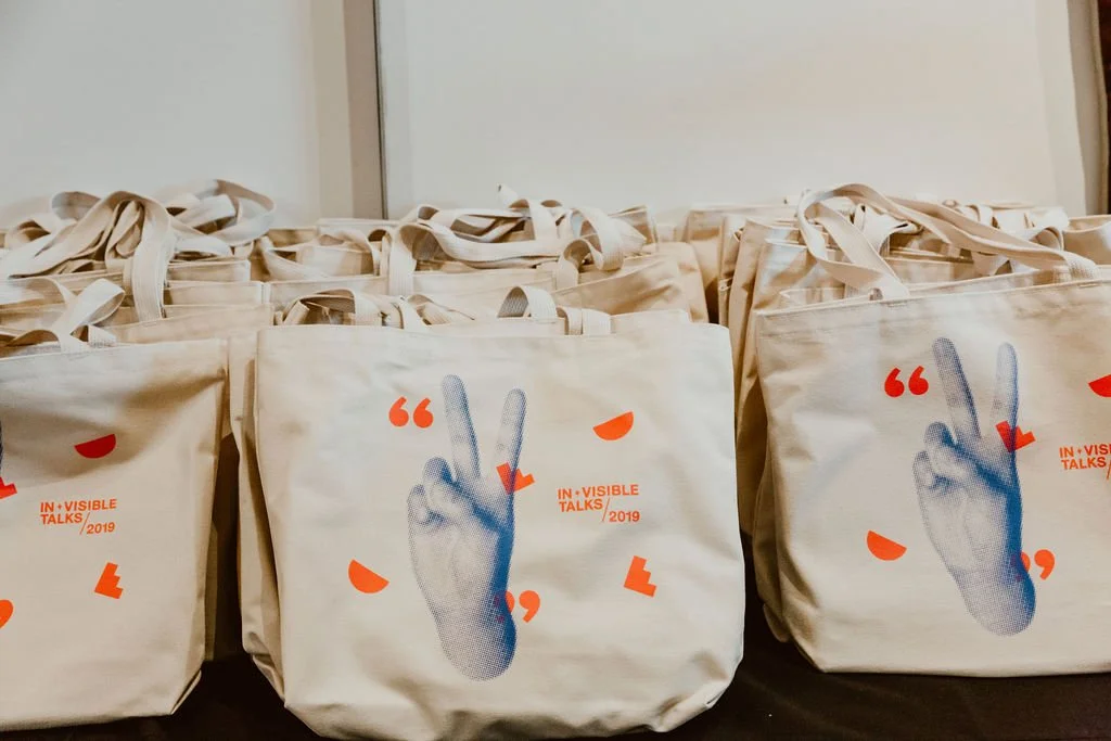 White tote bags with graphic design featuring a blue illustration of a hand making a peace sign and orange decorative elements, with text that says "IN VISIBLE TALKS/2019."