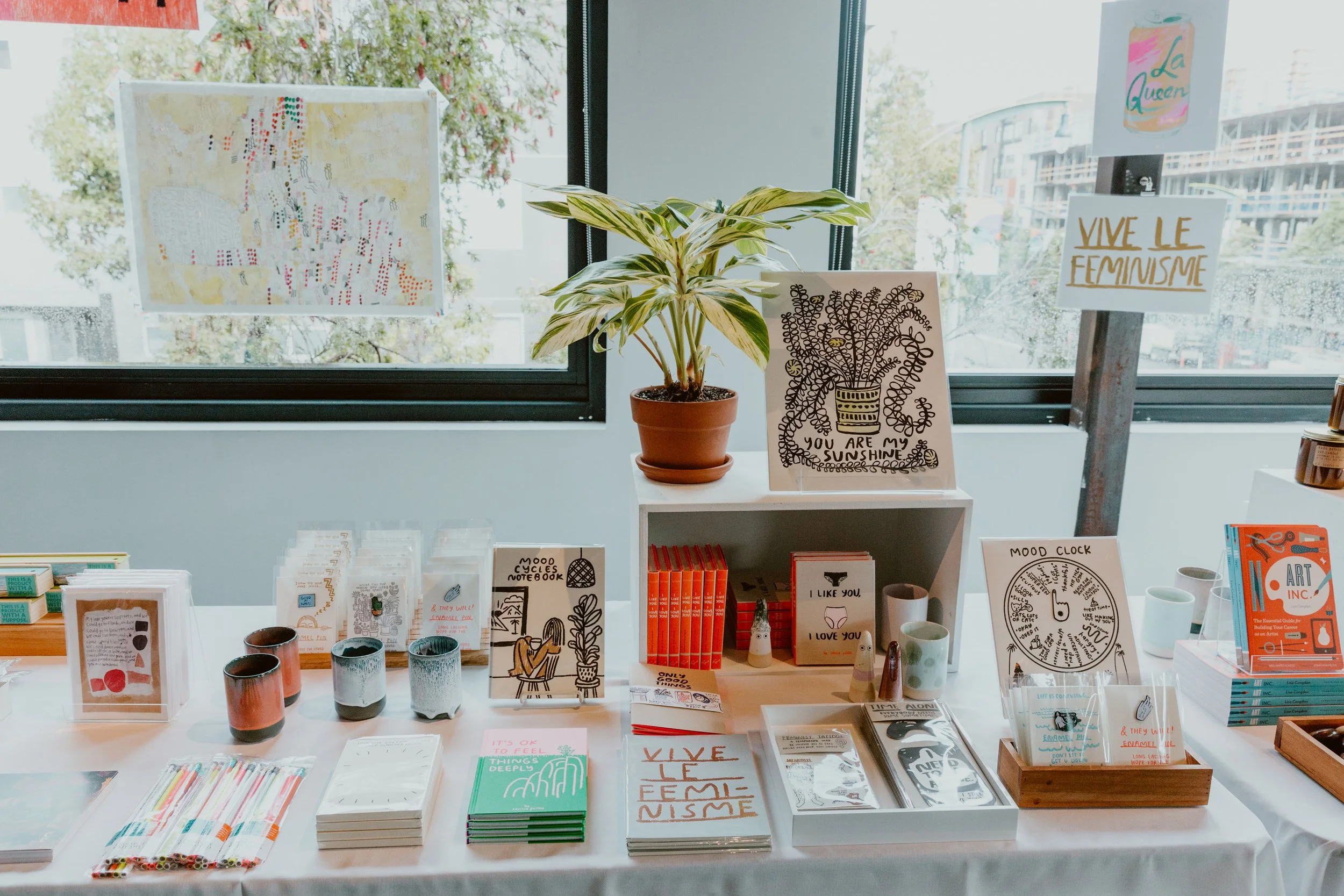 Display of various art and stationery items on a table in a store window, with handwritten signs and colorful notebooks, a potted plant, and abstract artwork in the background.