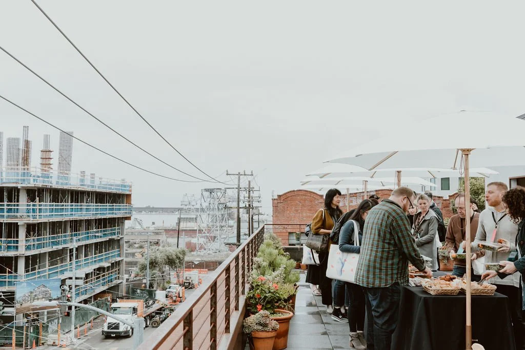 People standing in line at a buffet table on a rooftop terrace with umbrellas, overlooking an urban construction site with cranes and buildings.