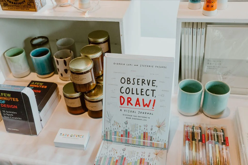 Display of ceramic mugs, jars, books, a visual journal titled "Observe, Collect, Draw!", and art supplies on a table.