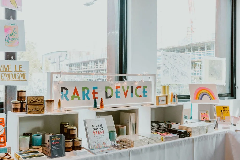 A display in a shop with a colorful sign reading 'RARE DEVICE', various books, art supplies, and decorative items, with large windows in the background.