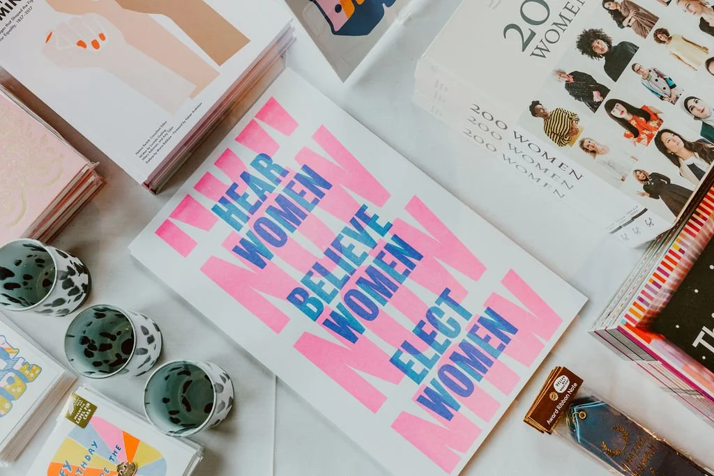 A table displaying pamphlets, books, and bookmarks, including a prominent pink and blue sign that reads "I HEAR WOMEN, I BELIEVE WOMEN, I EJECT WOMEN." There are also three black and white cow-patterned cups on the table.